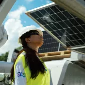 A person wearing a white hard hat, safety glasses, and a yellow safety vest is looking up at a solar panel. The person is standing outdoors with a partly cloudy sky in the background. Industrial equipment is visible around them.