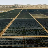 Aerial view of a vast solar farm with rows of solar panels stretching into the distance. The panels are arranged in a grid pattern on a flat, arid landscape. In the background, there are low, barren hills under a clear sky.