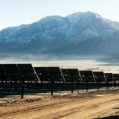 Rows of solar panels are installed on a dirt field, with a backdrop of snow-capped mountains under a clear blue sky.