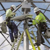 Two workers in safety gear and helmets install equipment on a metal structure. They are on ladders and handling cables, with a clear blue sky in the background.