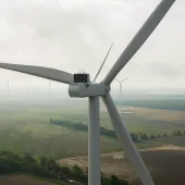Aerial view of a large wind turbine in the foreground with several other turbines scattered across a vast, flat landscape of fields and trees under a cloudy sky.