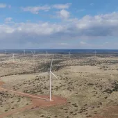 Aerial view of a wind farm in a vast, open landscape under a partly cloudy sky. Several wind turbines are scattered across the terrain, with dirt roads visible among sparse vegetation.