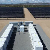 Aerial view of a large solar power plant in a desert landscape. Rows of solar panels stretch into the distance. In the foreground, there are several white electrical units and equipment on a gravel area. Power lines and mountains are visible on the horizon under a partly cloudy sky.