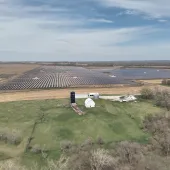 Aerial view of a large solar farm with rows of solar panels extending across a vast field. In the foreground, there is a farm with a white barn, silo, and a house surrounded by green grass and trees. The sky is partly cloudy.