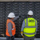 Two workers wearing hard hats and safety vests stand in front of a large battery storage system. One worker is pointing towards the batteries, while the other observes. The vest of the worker on the right has 'aes' written on it.