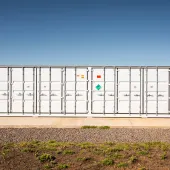 A large white industrial container with multiple doors and hazard labels is situated on a concrete platform. The background shows a clear blue sky and part of a solar panel array on the left. The foreground has gravel and patches of grass.