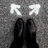 A person wearing black shoes stands on a gravel road in front of two white arrows painted on the ground, pointing in different directions, symbolizing a choice or decision.