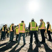 A group of people wearing hard hats and high-visibility vests stand in a circle on a construction site with mountains in the background. The sun shines brightly in the clear sky.