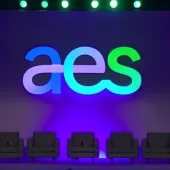 A stage setup with the large letters 'aes' illuminated in blue and green against a purple backdrop. Six empty gray chairs are lined up in front of the backdrop, and a podium with a microphone is on the right side.