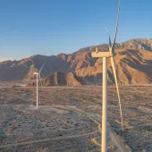 Aerial view of wind turbines in a desert landscape with mountains in the background. The turbines are surrounded by sparse vegetation under a clear blue sky during sunset, casting long shadows on the ground.