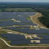 Aerial view of a large solar panel farm with rows of solar panels covering open fields. A small building and several parked cars are visible in the foreground. The landscape includes green fields, a river, and distant trees under a clear sky.