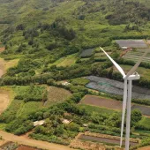 Aerial view of two large wind turbines situated among lush green farmland and hills. The landscape includes various agricultural plots with different shades of green, indicating diverse crops. Dirt roads wind through the area, and dense forested hills are visible in the background.