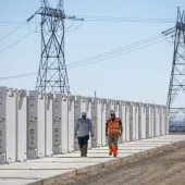 Two workers in safety gear walk along a row of large white battery storage units at a solar facility. High-voltage power lines and towers are visible in the background under a clear blue sky.