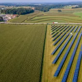 Aerial view of a large solar farm with rows of solar panels on the right side, adjacent to vast green agricultural fields on the left. In the background, a small rural community with buildings and roads is visible, surrounded by rolling hills and more farmland.