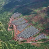 Aerial view of a large solar farm with multiple rows of solar panels surrounded by green vegetation and red soil. A winding road is visible on the left side, and the landscape is hilly with patches of sunlight and shadow.
