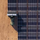 Aerial view of a large solar panel array on the right side, with a tractor and trailer carrying additional panels on the left, set on a dirt surface.