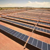 A large solar farm with rows of solar panels installed on a barren landscape. The panels are aligned in parallel rows under a clear blue sky. In the background, there is a view of distant mountains and some greenery.