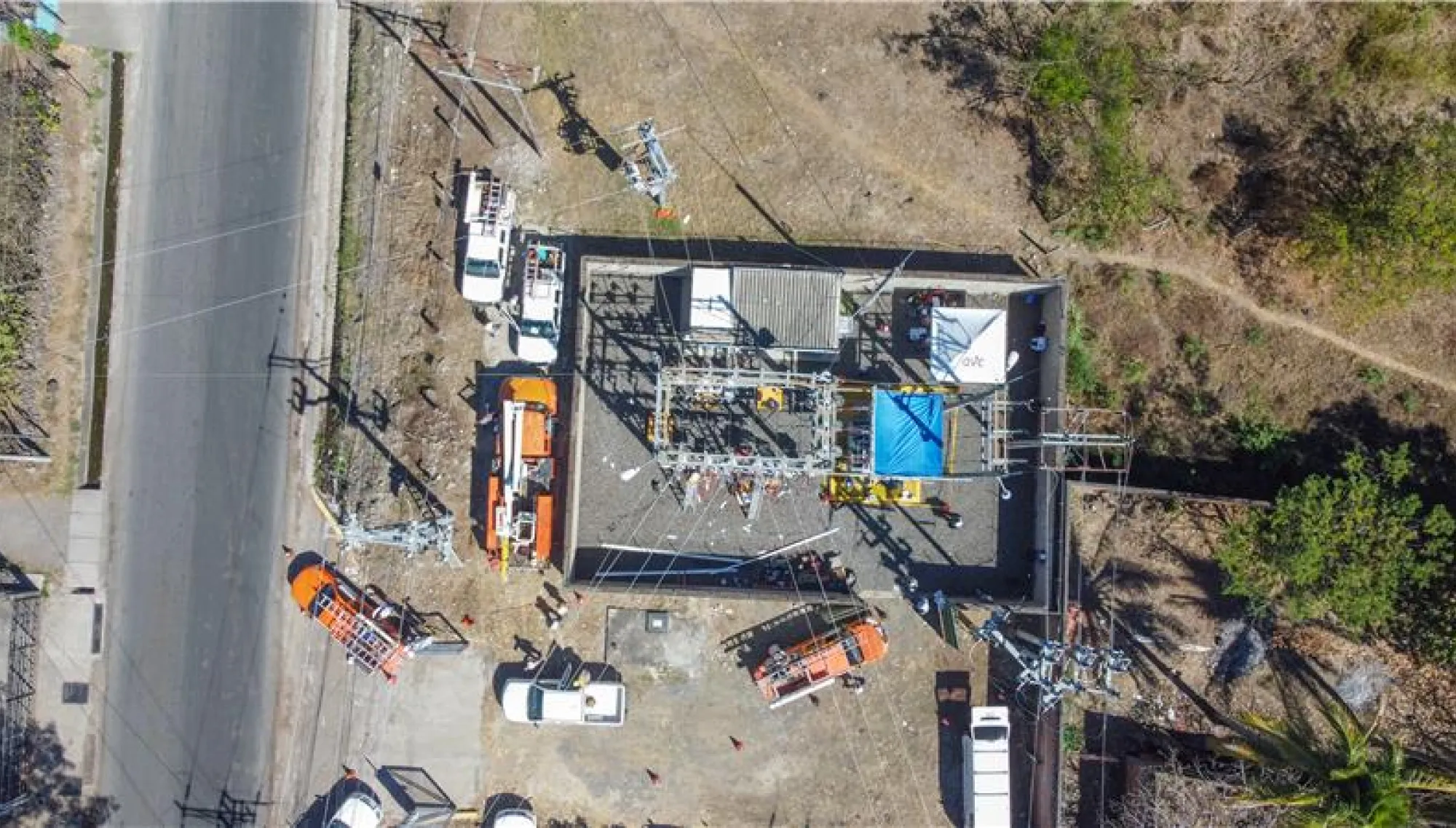 Aerial view of a power substation surrounded by several utility vehicles, including orange and white trucks, parked on a paved area. The substation is enclosed by a fence and contains electrical equipment. Nearby, there is a road on the left and grassy areas with trees on the right.