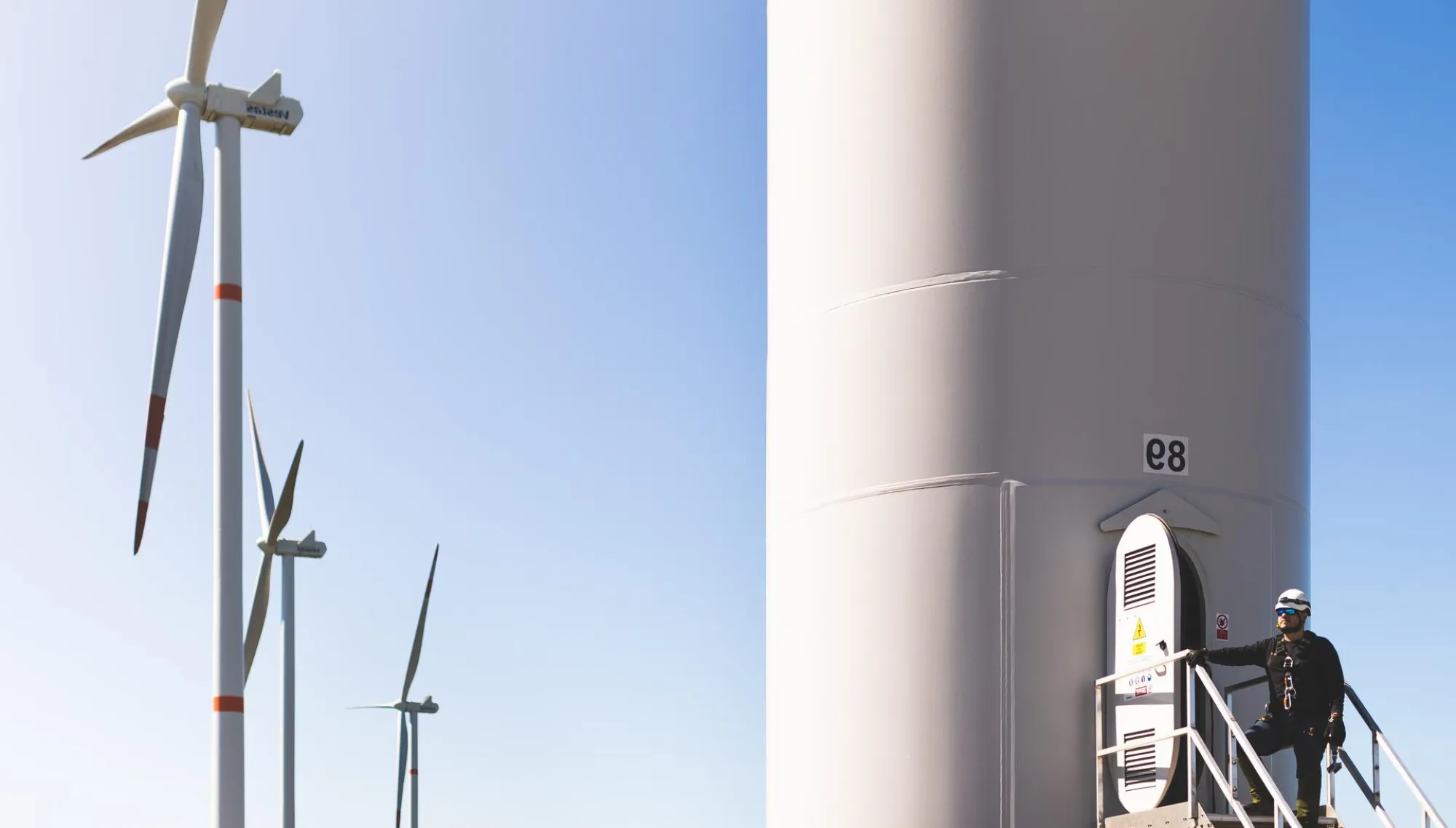 A person wearing safety gear stands on a platform attached to a large wind turbine tower labeled 'e8'. In the background, three more wind turbines are visible against a clear blue sky.