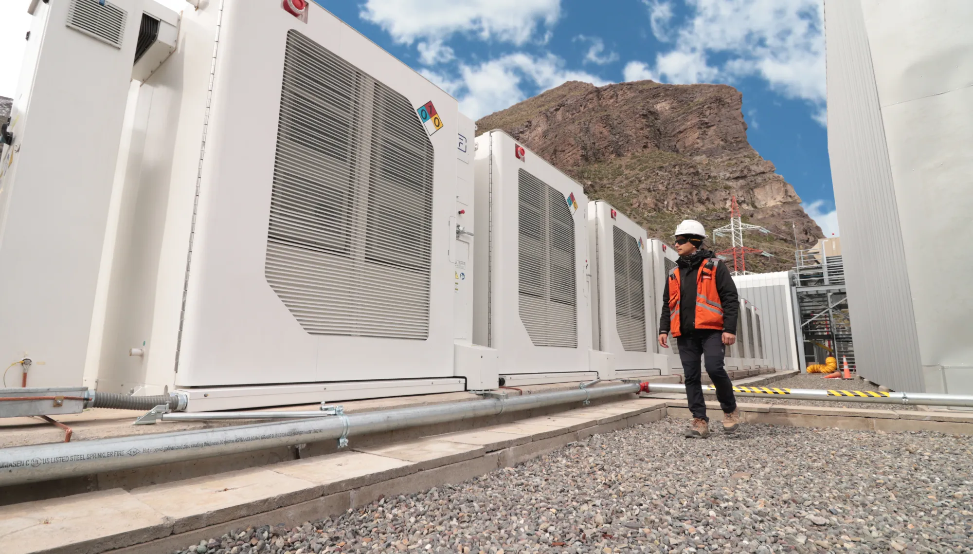 A person wearing a hard hat and orange safety vest walks beside large industrial cooling units in a mountainous area. The sky is clear with a few clouds.