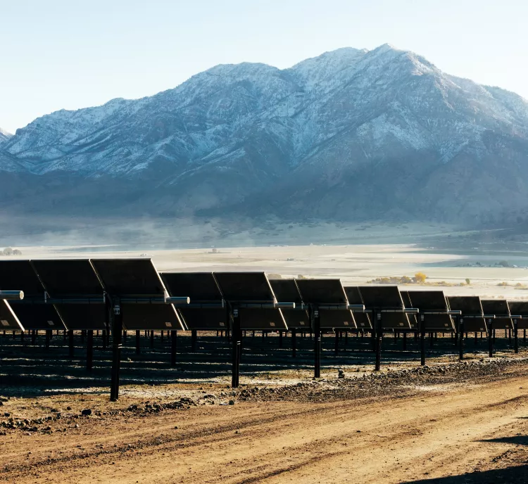 Rows of solar panels are set up in a barren landscape with a dirt road in the foreground. Snow-capped mountains rise in the background under a clear blue sky.