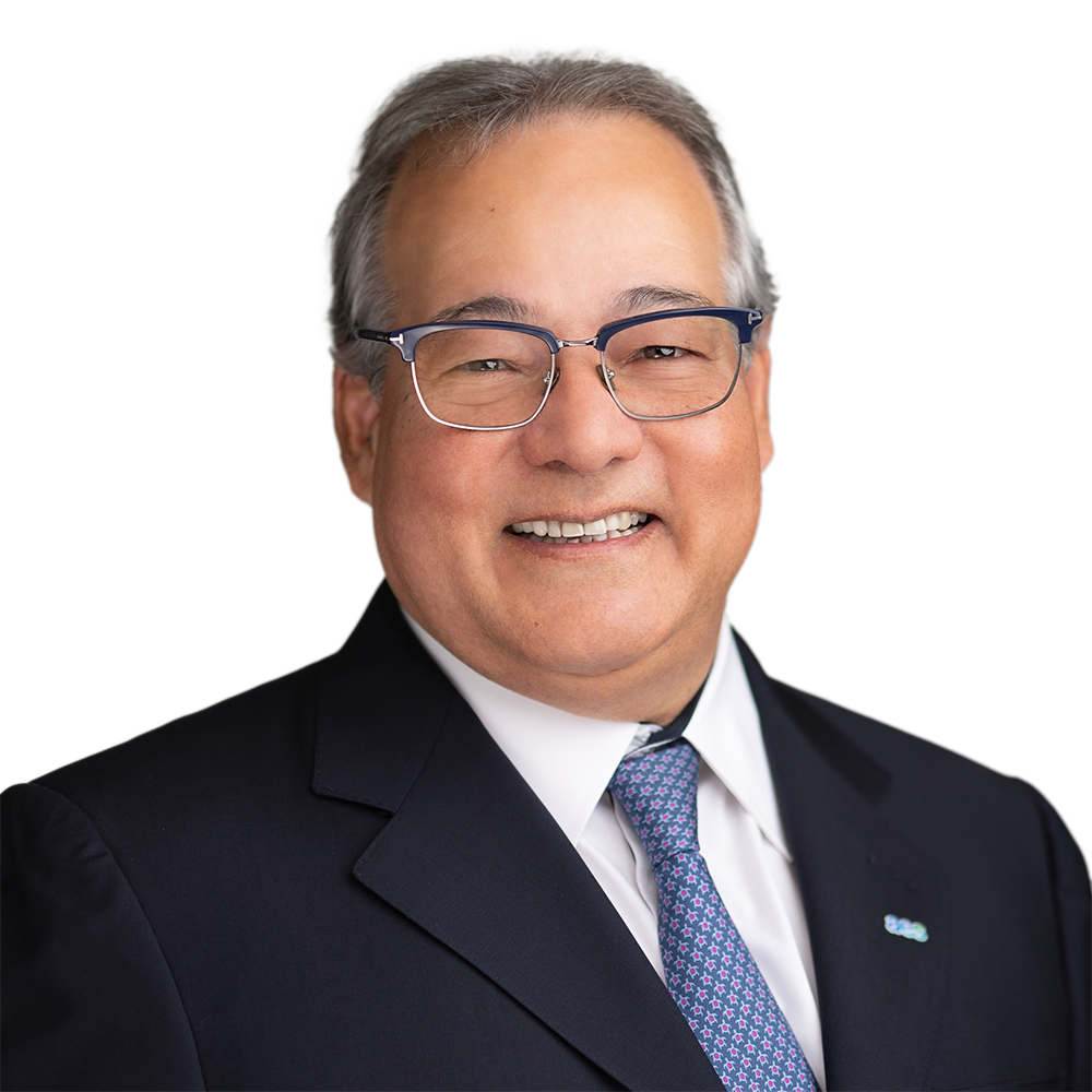 A man with short gray hair and glasses is smiling, wearing a dark suit, white shirt, and patterned tie.