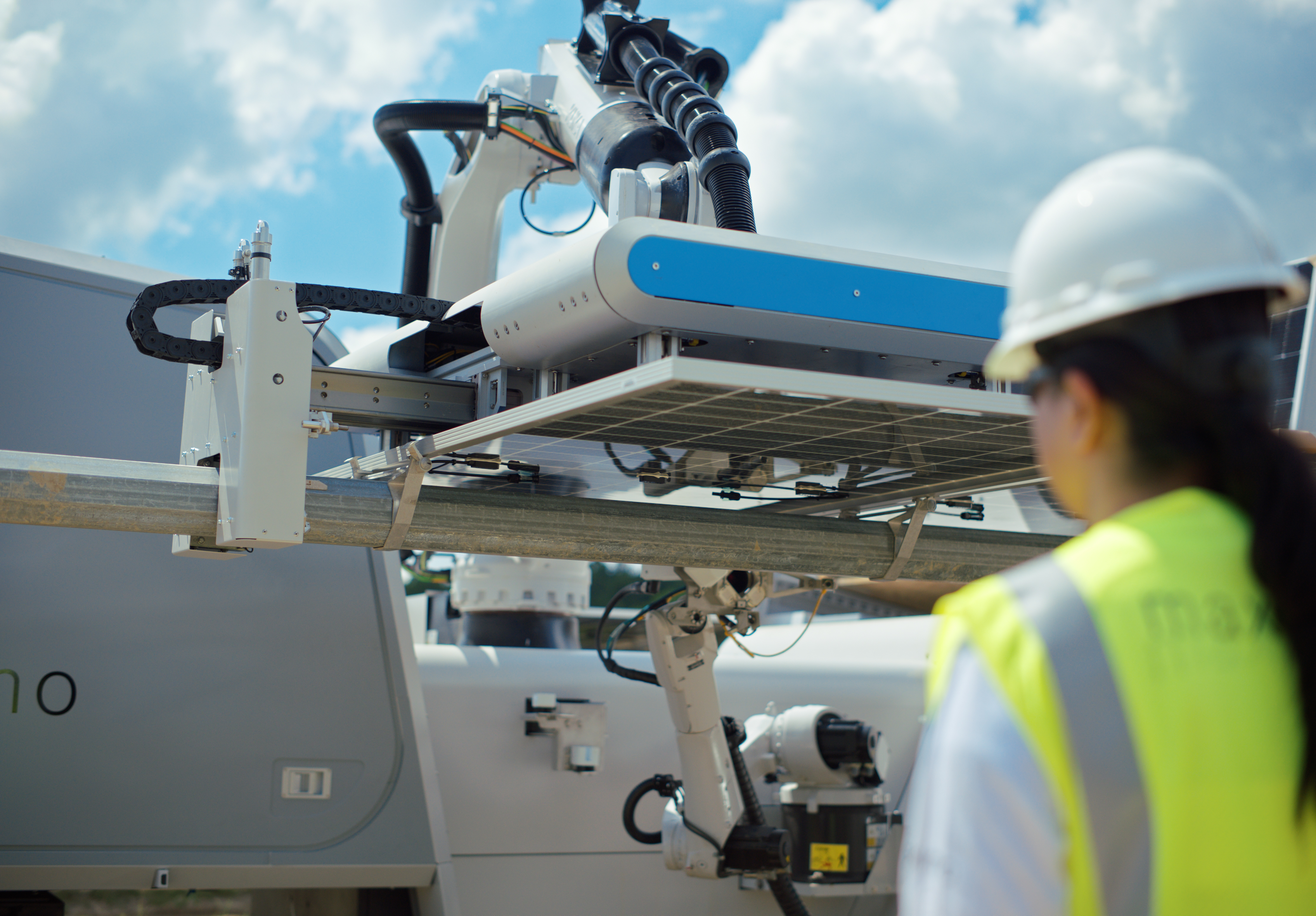 A worker in a yellow safety vest and helmet observes a robotic arm installing a solar panel under a blue sky.