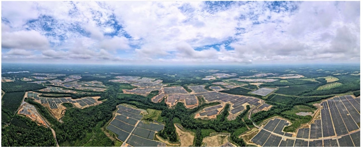 Aerial view of a vast solar farm with numerous solar panels spread across a landscape of green forests and open fields under a partly cloudy sky.