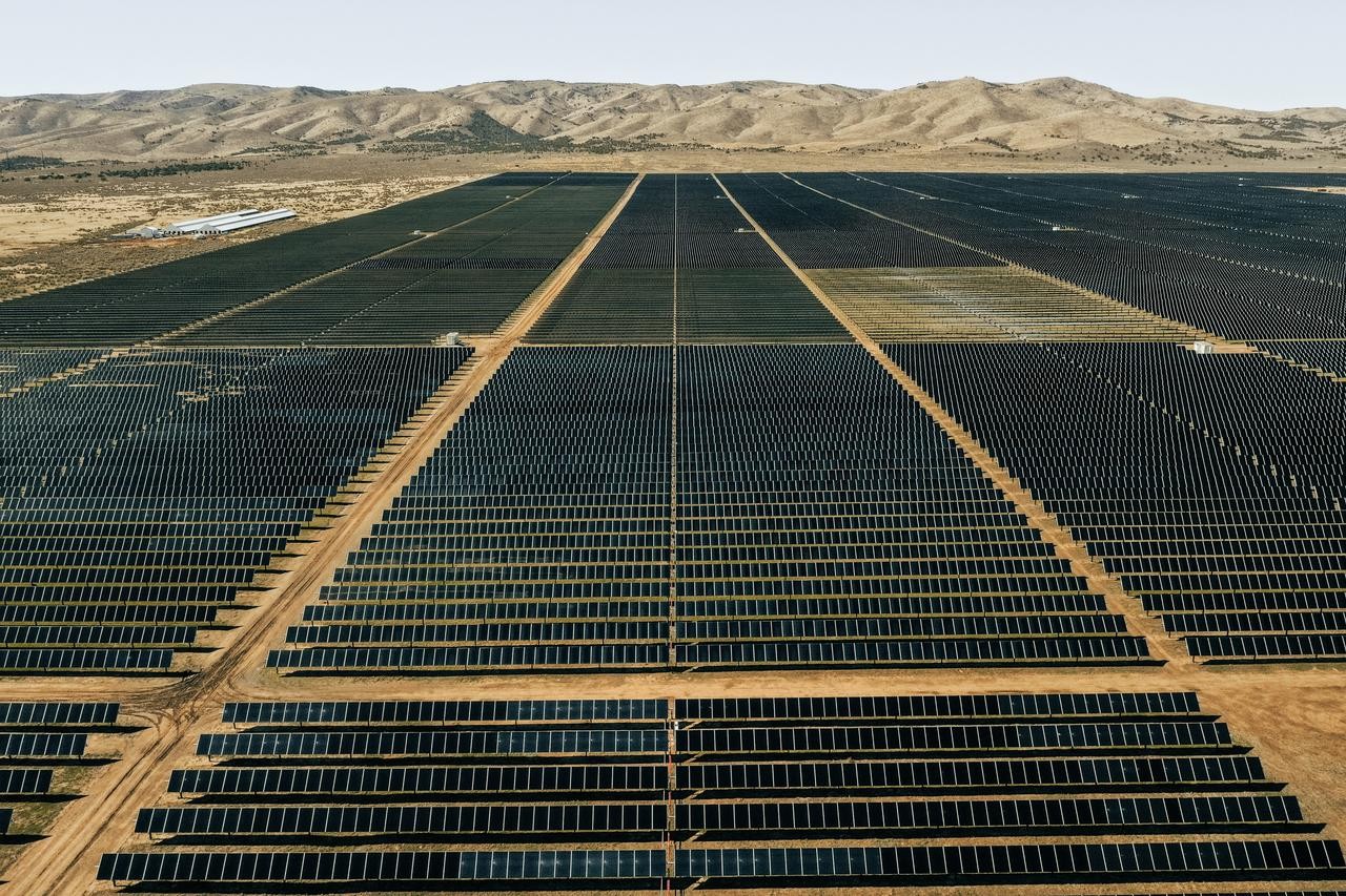 Aerial view of an extensive solar farm with rows of solar panels stretching into the distance, set against a backdrop of arid hills and a clear sky.