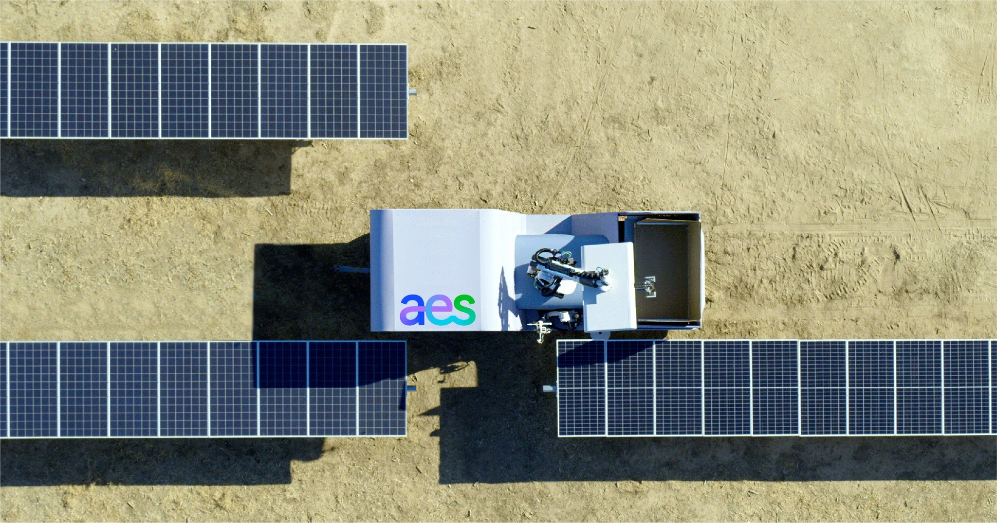 Aerial view of solar panels on a sandy surface with a robotic cleaning device labeled 'aes' positioned between them.