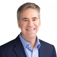 A man with short gray hair wearing a blue suit jacket and light blue shirt smiles against a white background.