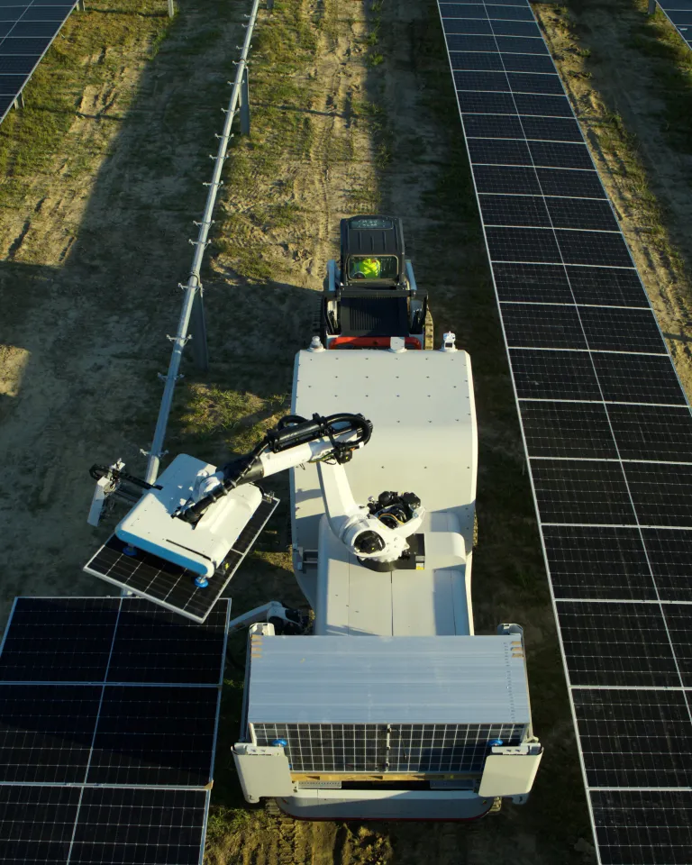 Aerial view of a Maximo robotic machine cleaning solar panels in a field. The machine is positioned between rows of solar panels, using an arm to clean the surface. The ground is partially covered with grass and dirt.