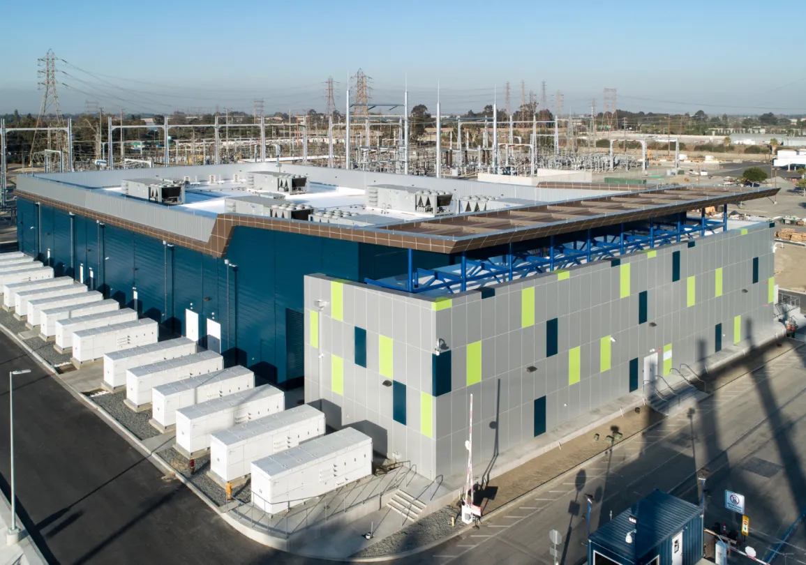 Aerial view of a modern industrial facility with a blue and gray building surrounded by electrical infrastructure and multiple white storage units.