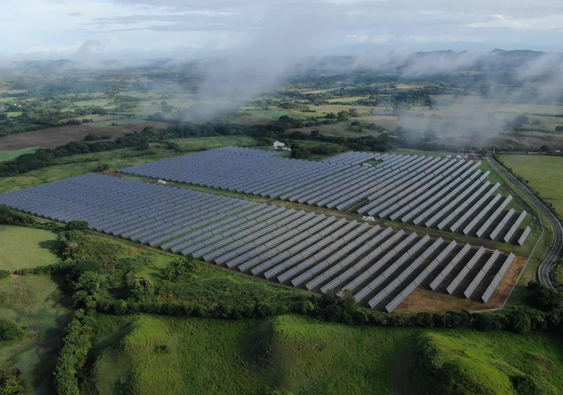 Aerial view of a large solar farm with rows of solar panels surrounded by lush green fields and distant hills under a cloudy sky.
