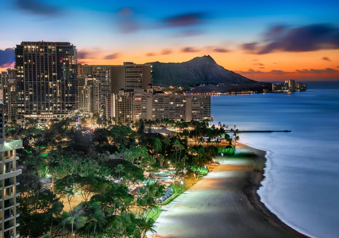 Evening view of Waikiki Beach with illuminated city buildings and Diamond Head in the background, under a colorful sunset sky.
