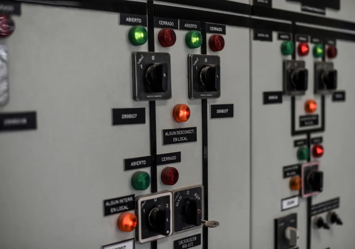 Close-up of an industrial control panel with various switches, dials, and indicator lights. The panel features red and green lights labeled 'Cerrado' and 'Abierto,' along with black switches and a key inserted in one of the switches. Labels in Spanish such as 'Algun Desconect. En Local' are visible.