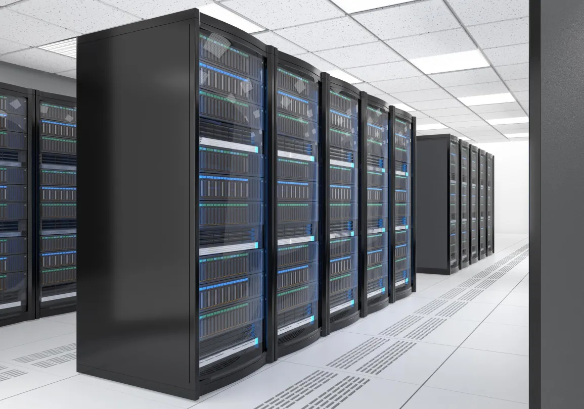 A modern data center with rows of black server racks filled with equipment. The floor is white with ventilation panels, and the ceiling is lined with fluorescent lights. The room is well-lit and organized, with a clear aisle between the server racks.