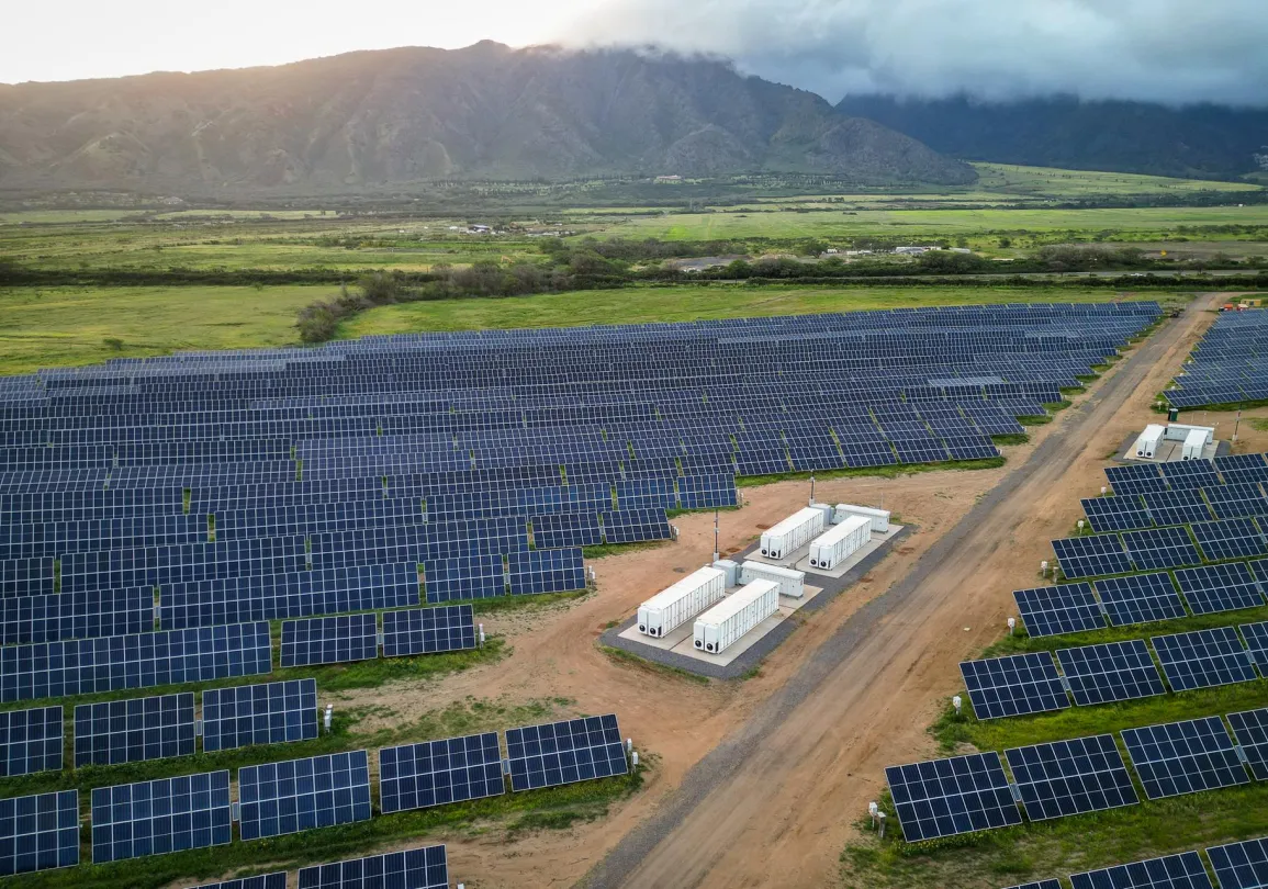 Aerial view of a large solar farm with numerous rows of solar panels. In the center, several white utility buildings are visible. The landscape includes green fields and distant mountains under a cloudy sky.