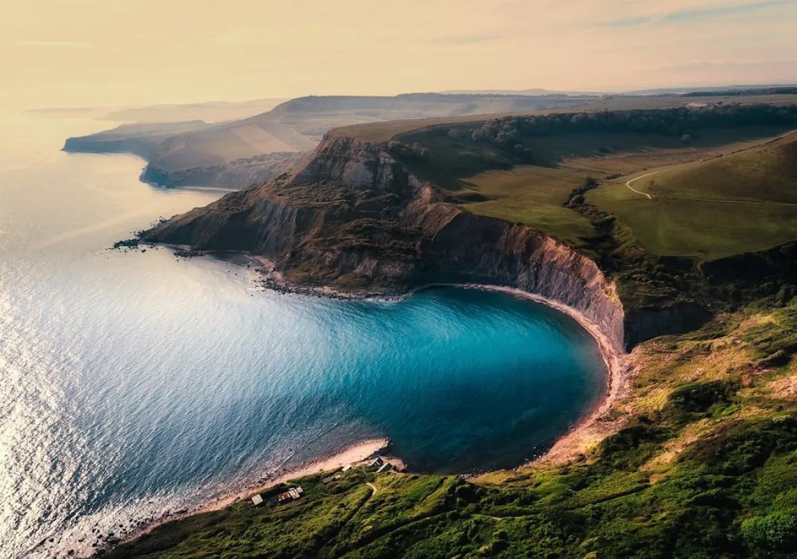 Aerial view of a coastal landscape with steep cliffs and a curved bay with turquoise water. The cliffs are covered in green vegetation, and the sea reflects the light of the setting sun.
