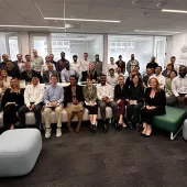 A diverse group of people, approximately 40 individuals, are gathered in an office setting. They are smiling and seated on modern furniture, including sofas and ottomans, with some standing at the back. The room has a contemporary design with glass walls and framed posters on the wall.