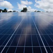 Close-up of a solar panel array under a blue sky with scattered clouds. The panels are aligned in rows, reflecting the sky and clouds above. Trees are visible in the distant background.