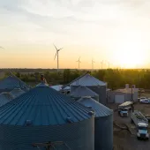 A rural landscape at sunset featuring several large metal silos in the foreground, wind turbines in the background, and a few vehicles parked near a building.