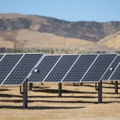 Rows of solar panels installed on a dry, brown field with hills in the background under a clear blue sky.