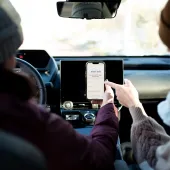 Two people in a car, one holding a smartphone displaying a screen with the text 'MOTOR' and other visible details. The person on the right is pointing at the phone screen. Both are wearing winter clothing.