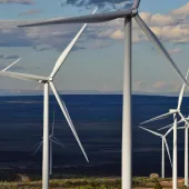 A series of wind turbines with large blades stand in an open landscape under a partly cloudy sky. The turbines are aligned in a row, stretching into the distance, with hills visible on the horizon.