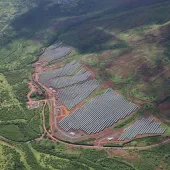 Aerial view of a large solar farm with rows of solar panels surrounded by lush green hills and a winding road.