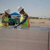 Two construction workers in safety vests and helmets inspect solar panels stacked on a pallet at a solar farm. A tractor and more solar panels are visible in the background under a clear sky.