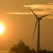 A wind turbine stands tall against a golden sunset sky, with the sun shining brightly near the horizon. Silhouettes of trees are visible at the bottom of the image.