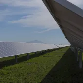 Rows of solar panels on metal stands in a field of green grass under a blue sky with scattered clouds. A mountain range is visible in the distant background.