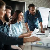 Four people in a modern office setting, sitting and standing around a computer screen, smiling and discussing. One person holds papers, and a whiteboard is visible in the background.